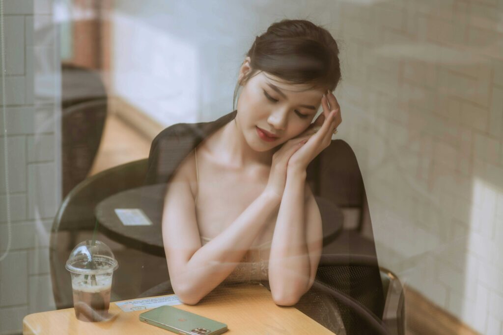Woman resting her head on her hands at a cafe table, looking down thoughtfully with a iced drink and phone nearby on the table.