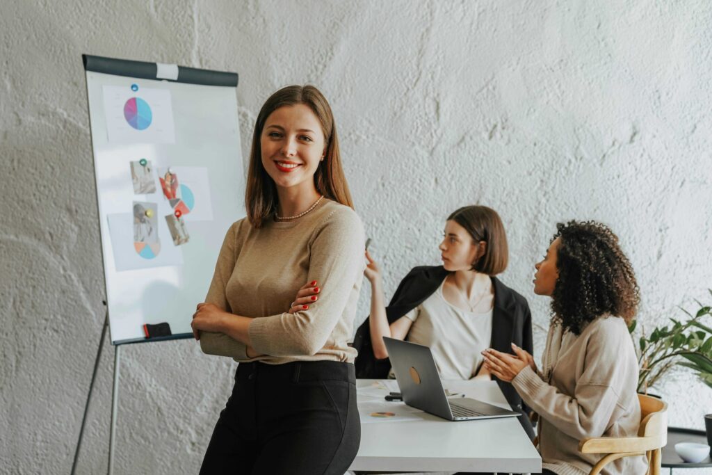 Confident woman stands with arms crossed in front of a whiteboard as colleagues discuss charts in a bright office.