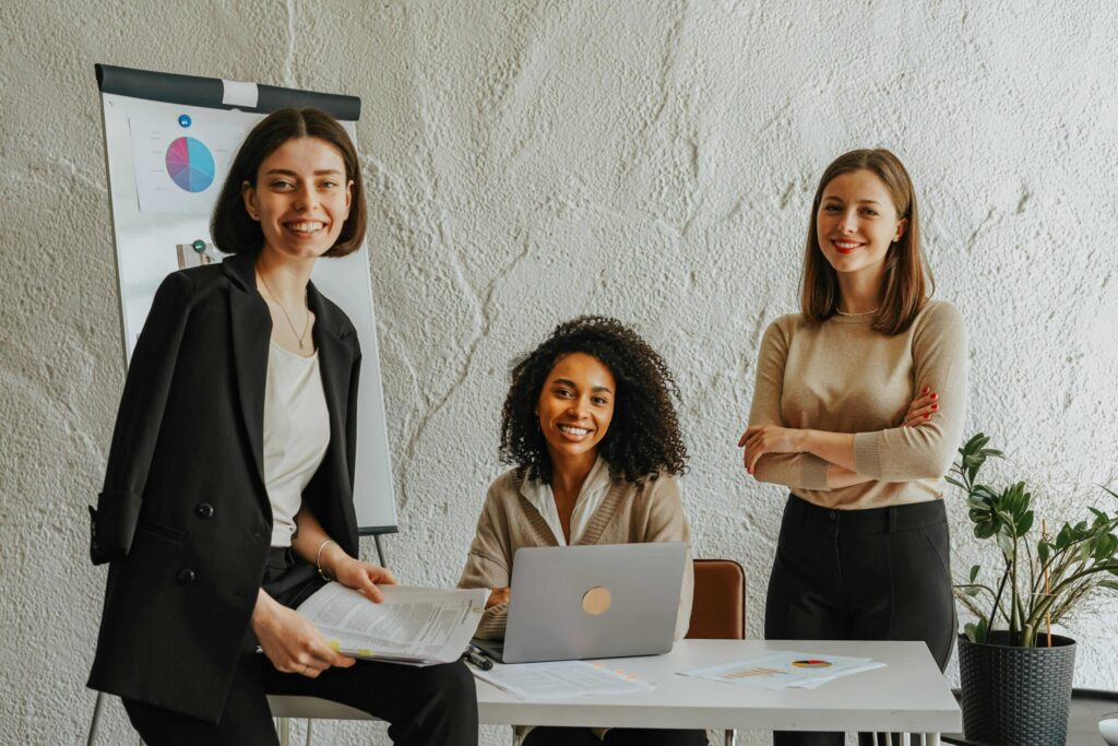 Three diverse professionals in a meeting: two women standing and one seated at a table with a laptop and documents.