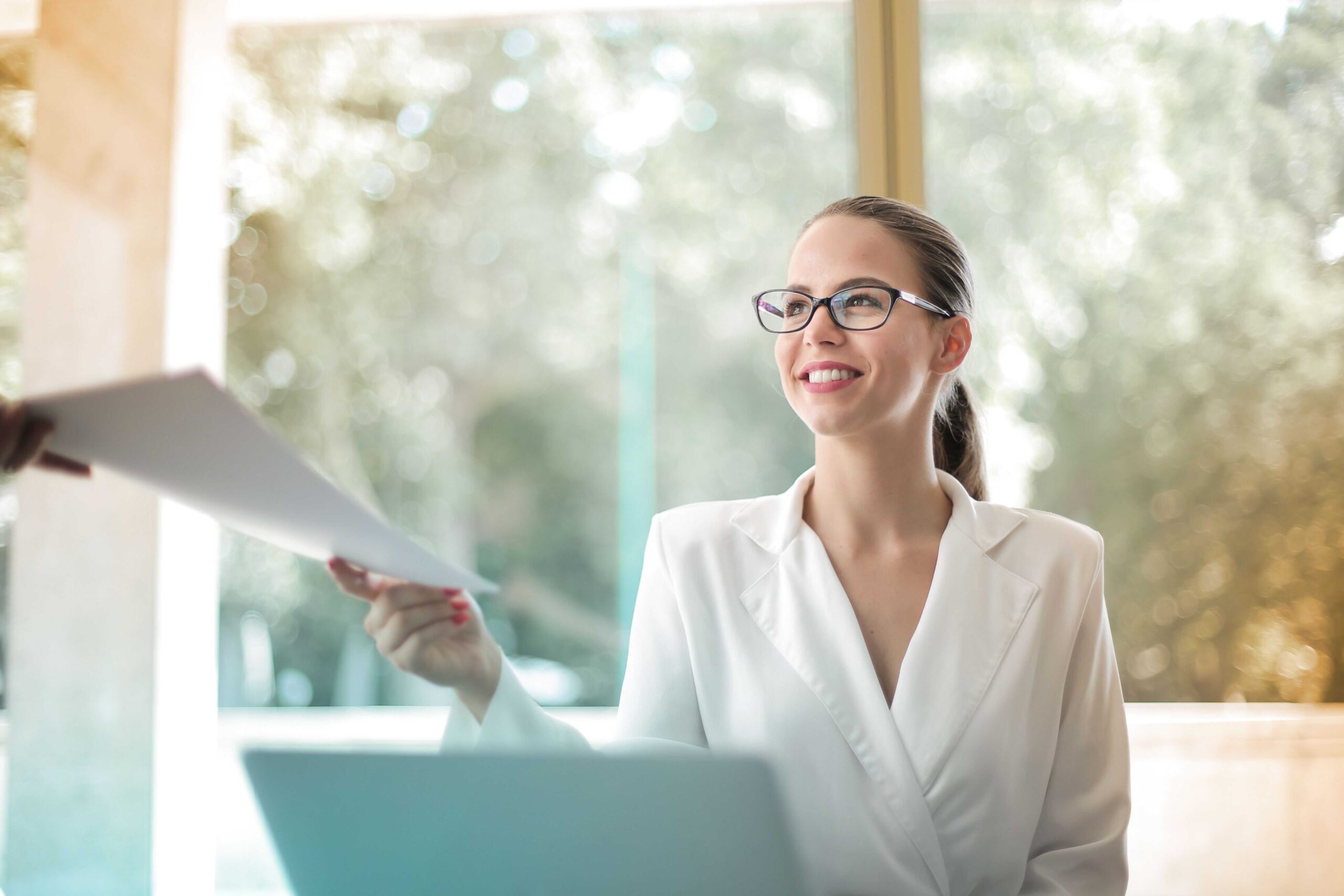 Professional woman in a white blazer hands a document to someone off-camera, smiling in a bright office setting