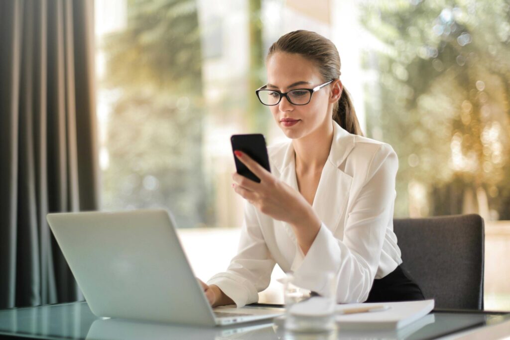 Businesswoman wearing glasses in a white blazer sits at a glass desk, looking at her smartphone with a laptop nearby in a bright office setting.