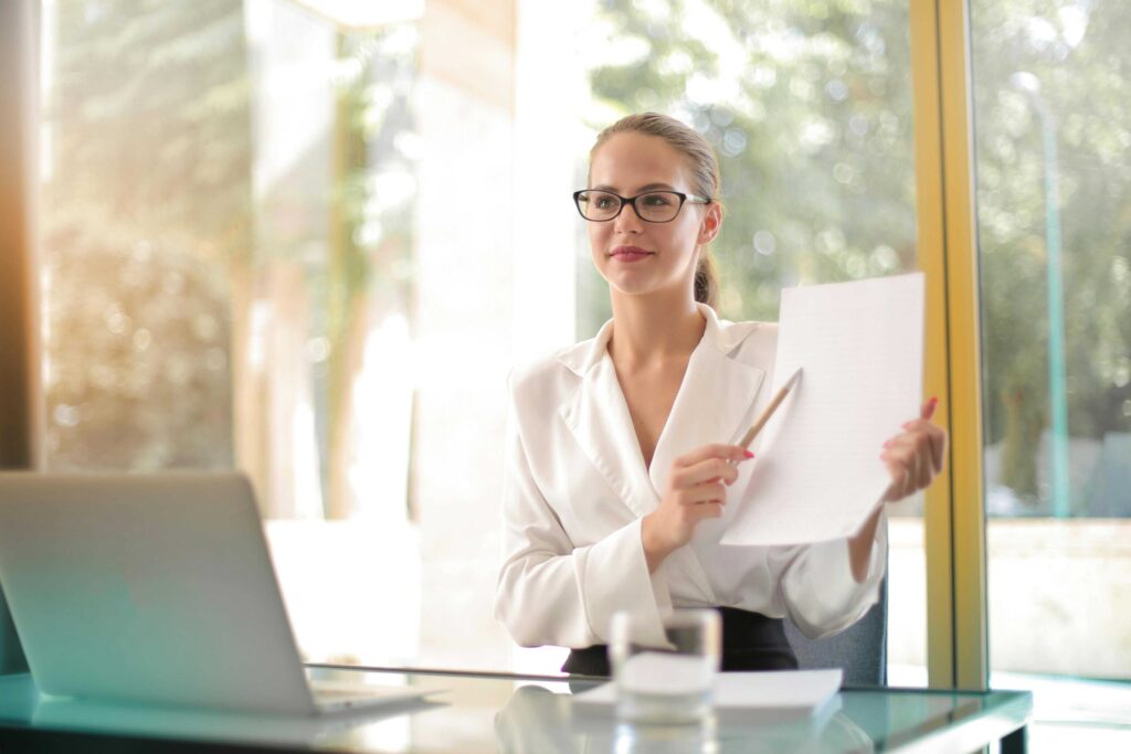 Professional woman wearing glasses and a white blazer holds papers in a bright, glass-walled office.