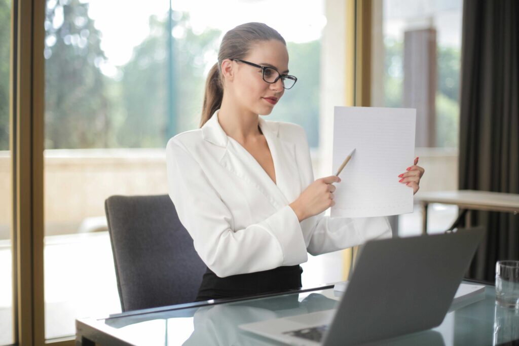 Businesswoman in a white blazer reviews a document at a glass desk with a laptop nearby, in an office setting, wearing glasses and holding a pencil.