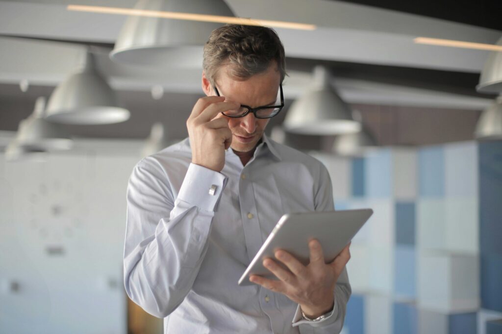 Man in a light shirt adjusts his glasses while reading a tablet in a modern office.