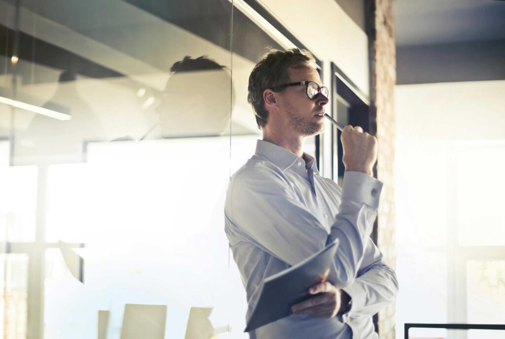 Man in a light blue shirt and glasses, standing beside a glass wall, holding a pen to his lips in a contemplative pose in an office.