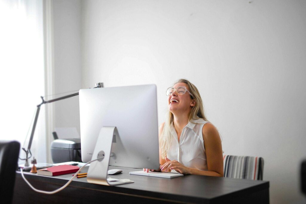 Woman with blonde hair and glasses sitting at a desk, smiling as she types on a desktop computer in a bright office.