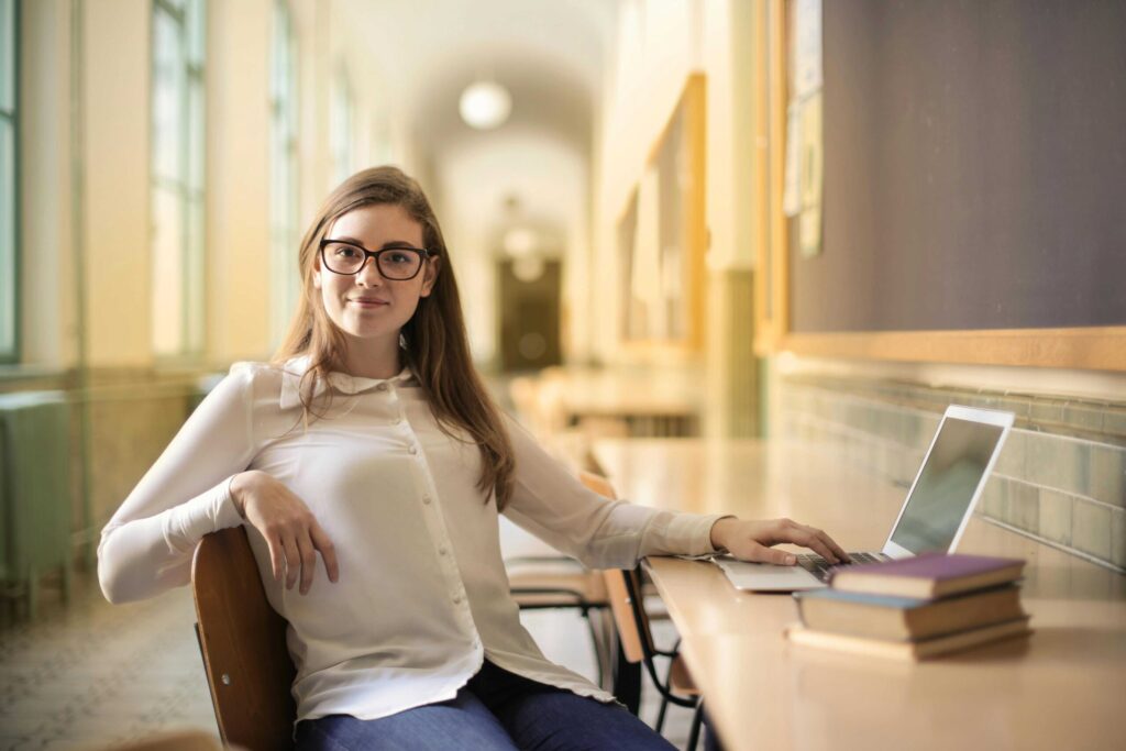 Student with glasses sits at a desk in a library, using a laptop with a stack of books nearby in a quiet study area.