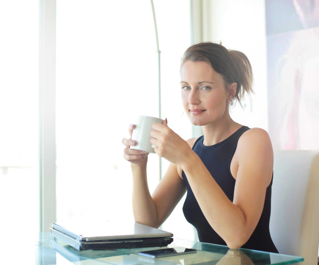 Woman in a black sleeveless top holding a mug near a laptop on a glass table by a bright window area.