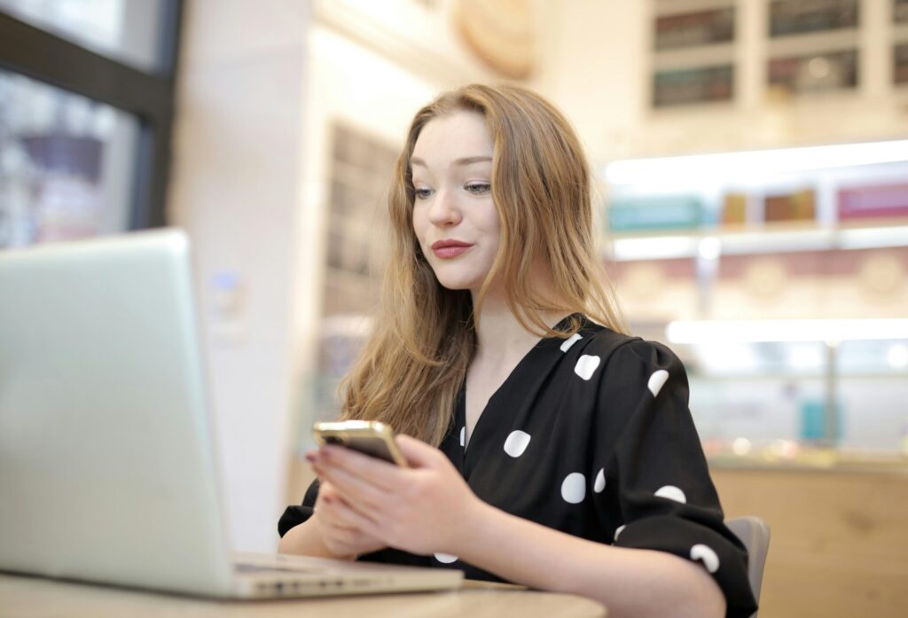 Woman with long hair looking at a smartphone while sitting at a desk with a laptop in a bright cafe or workspace.