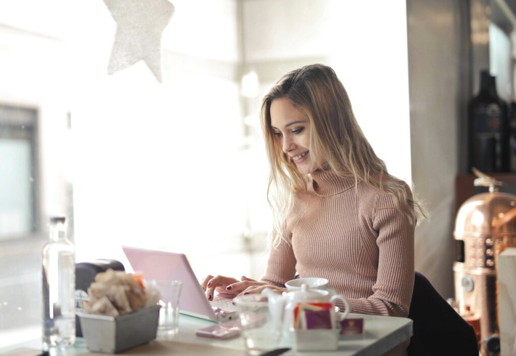 Woman in a pink ribbed sweater sits at a cafe table, typing on a pink laptop with coffee cups nearby.