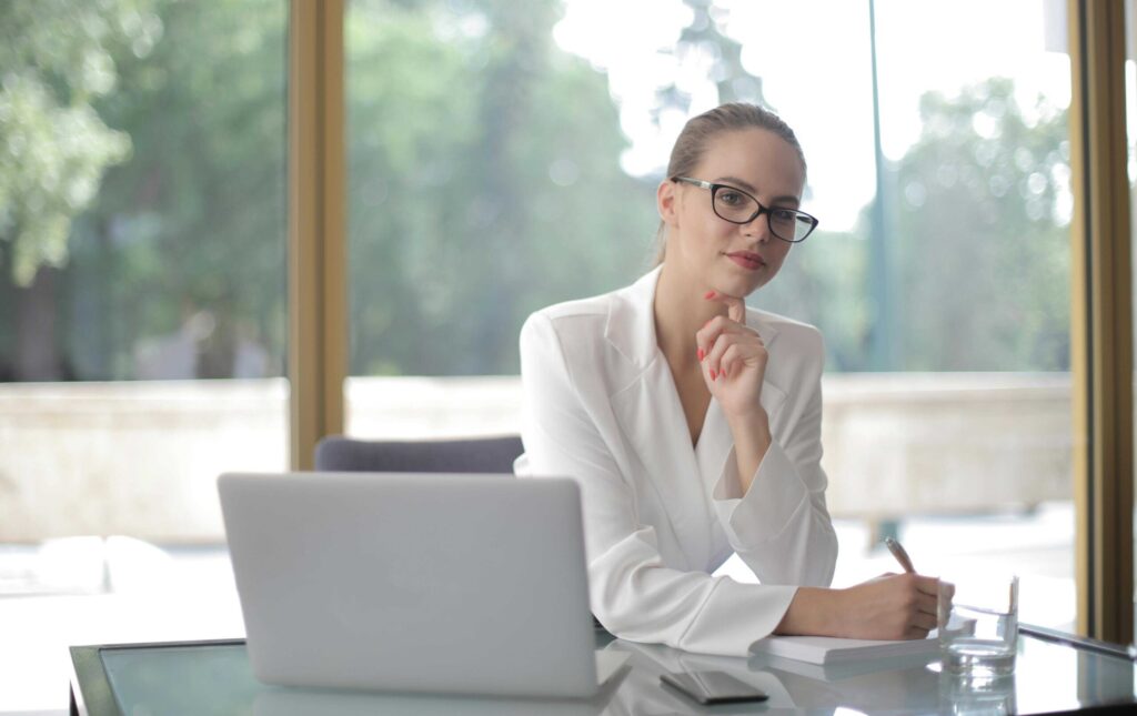 Professional woman in a white blazer and glasses sits at a glass desk with a laptop, resting her chin on her hand as she thinks.