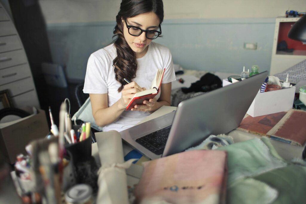 Woman with glasses writes in a small red notebook at a cluttered desk with a laptop open in front of her.