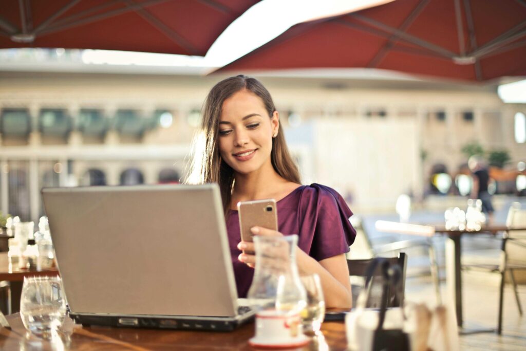 Woman seated at an outdoor cafe using a laptop and looking at her smartphone, with drinks on the table under red umbrellas.
