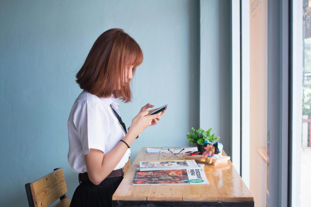 A young woman wearing a white shirt and black skirt stands at a wooden table, looking at her smartphone, by a blue wall and window.