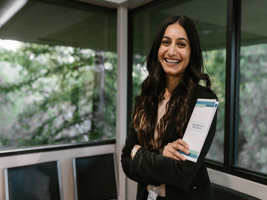Smiling woman in a blazer holding a conference program booklet in a glass-walled hallway