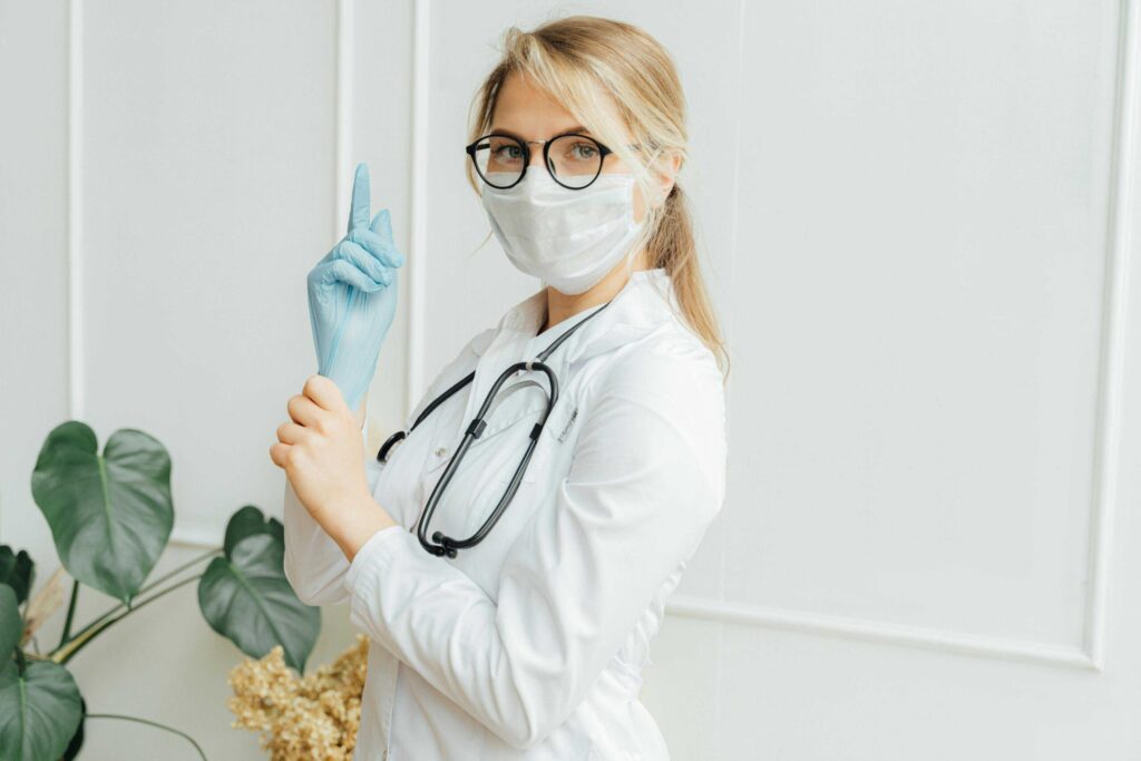 Female healthcare worker in a white coat, mask, and stethoscope raises one gloved finger, indoors with plants nearby.