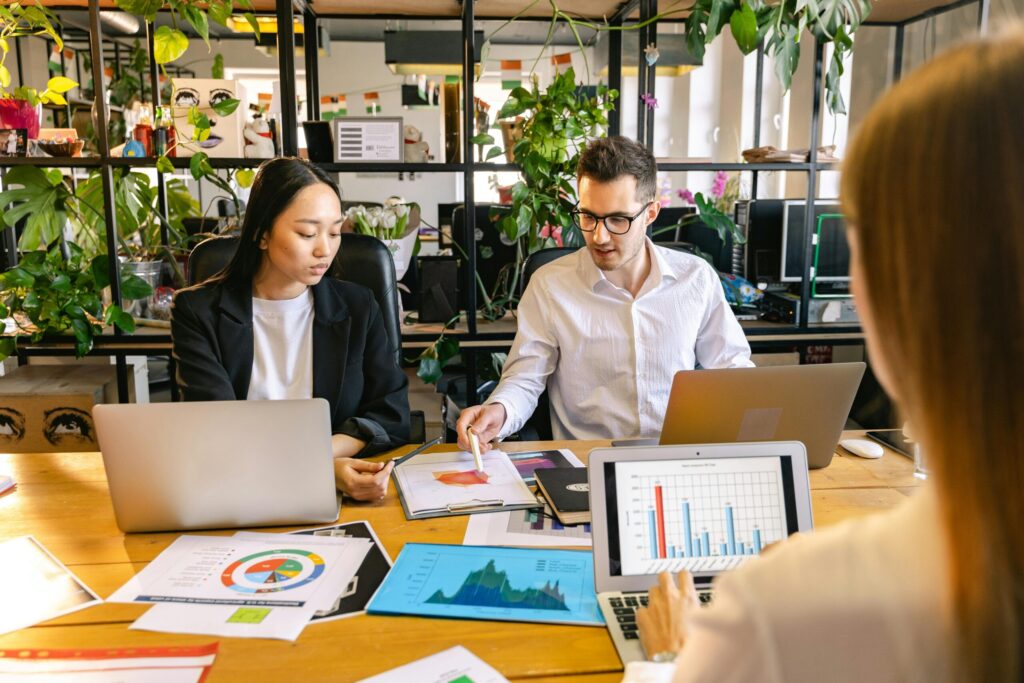 Three colleagues collaborate at a wooden table with laptops and printed charts in a bright, plant-filled office space