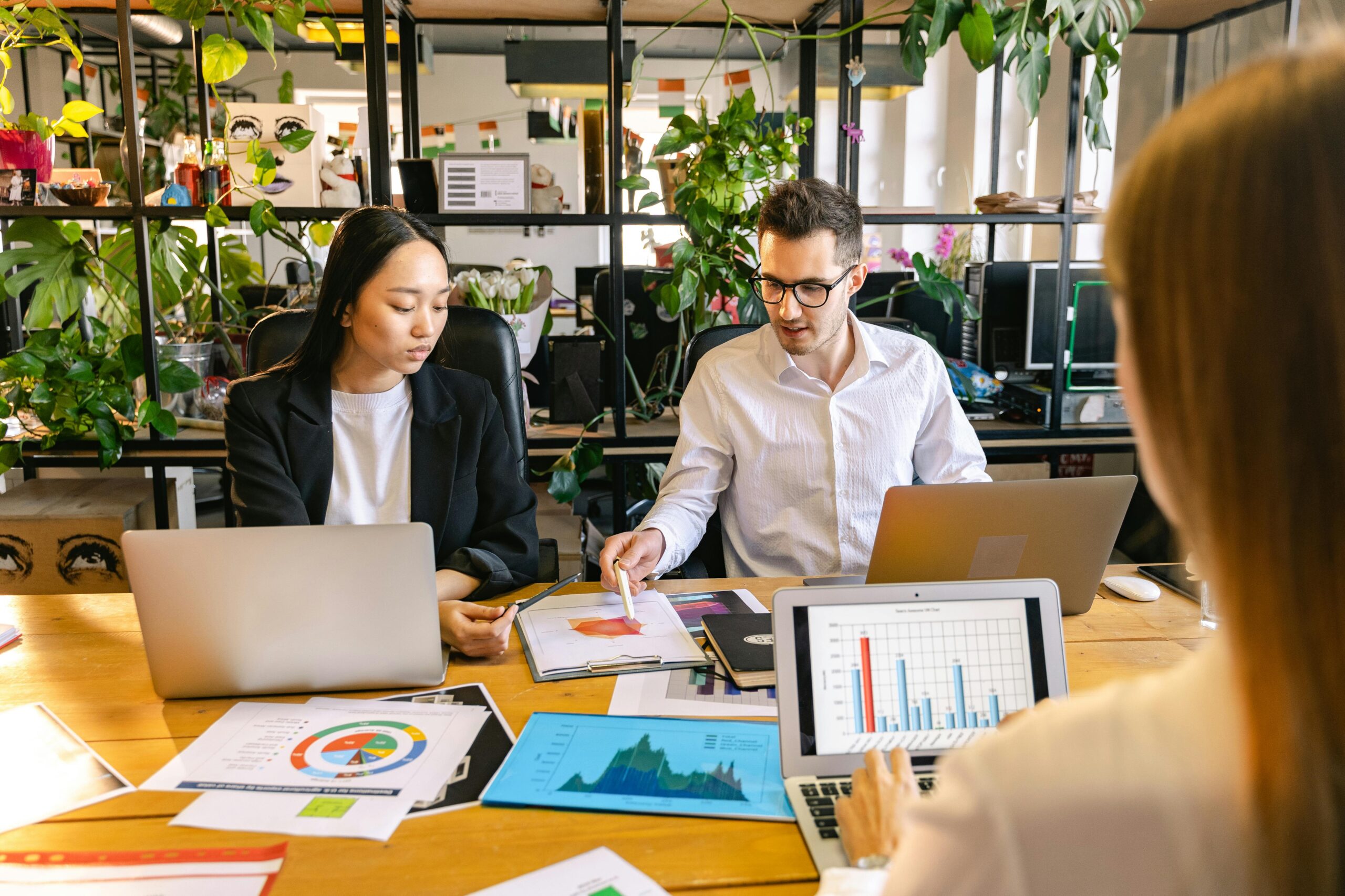 Three colleagues collaborate at a wooden table with laptops and printed charts in a bright, plant-filled office space