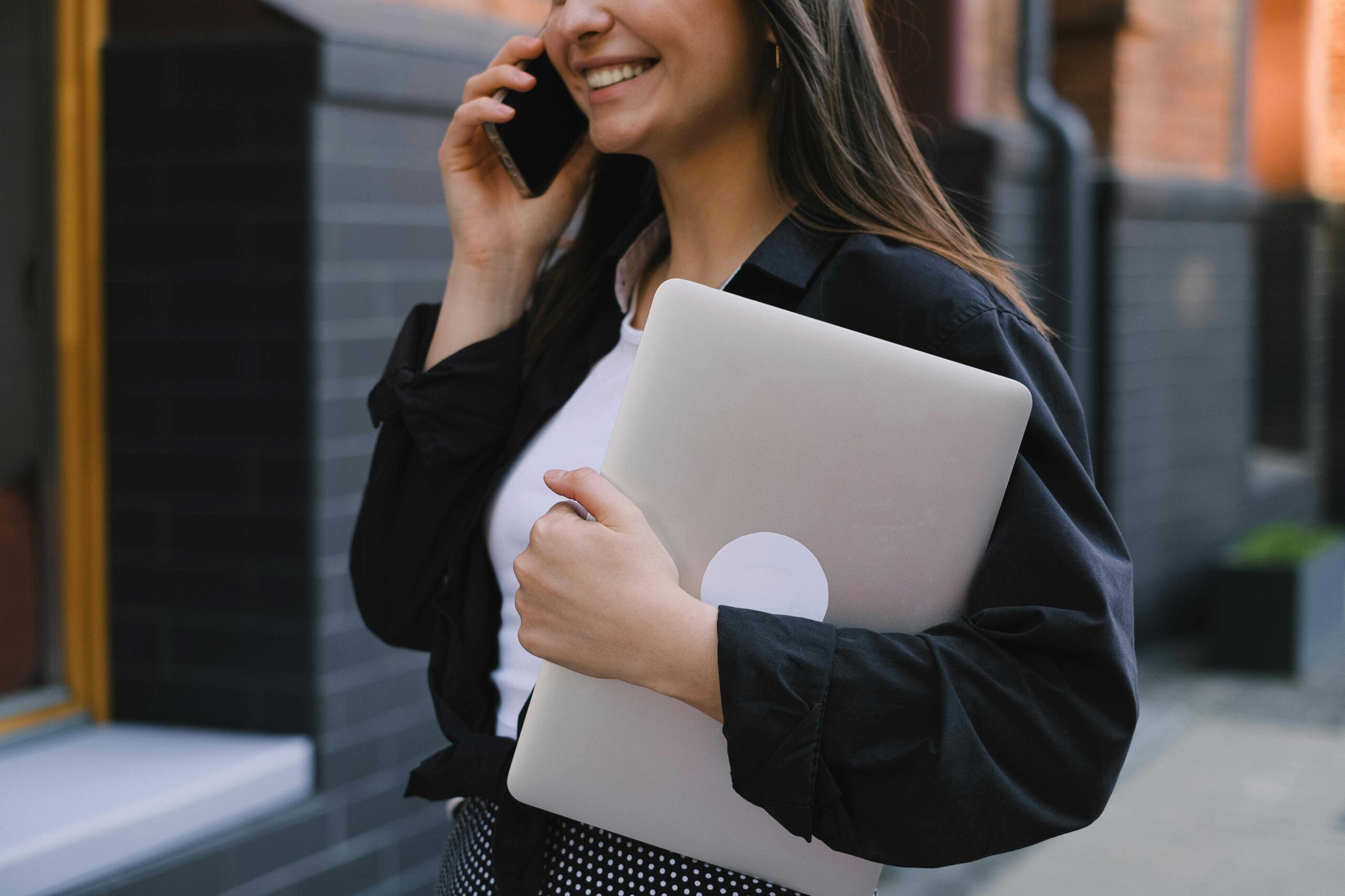 Woman smiling while talking on a smartphone, holding a silver laptop under her arm outdoors edge of a building behind her