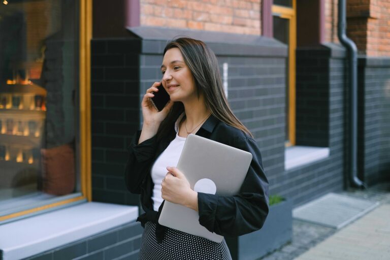 Professional woman in a black jacket talks on her phone while holding a silver laptop outdoors near a brick building.