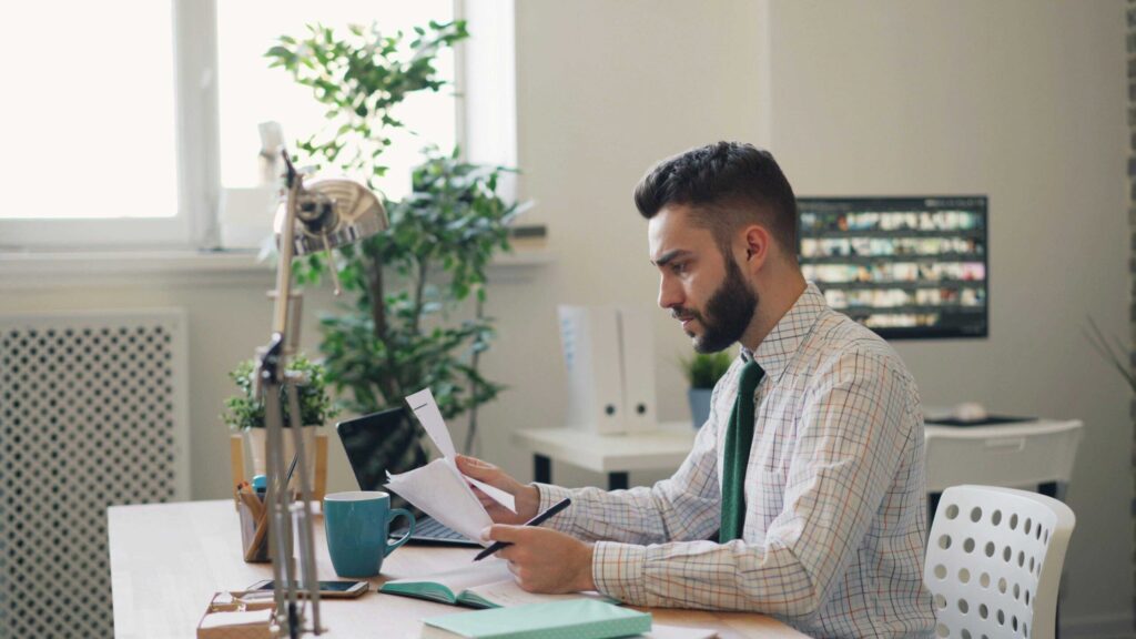 Bearded man in a checkered shirt reviews documents at a bright office desk with plants nearby.