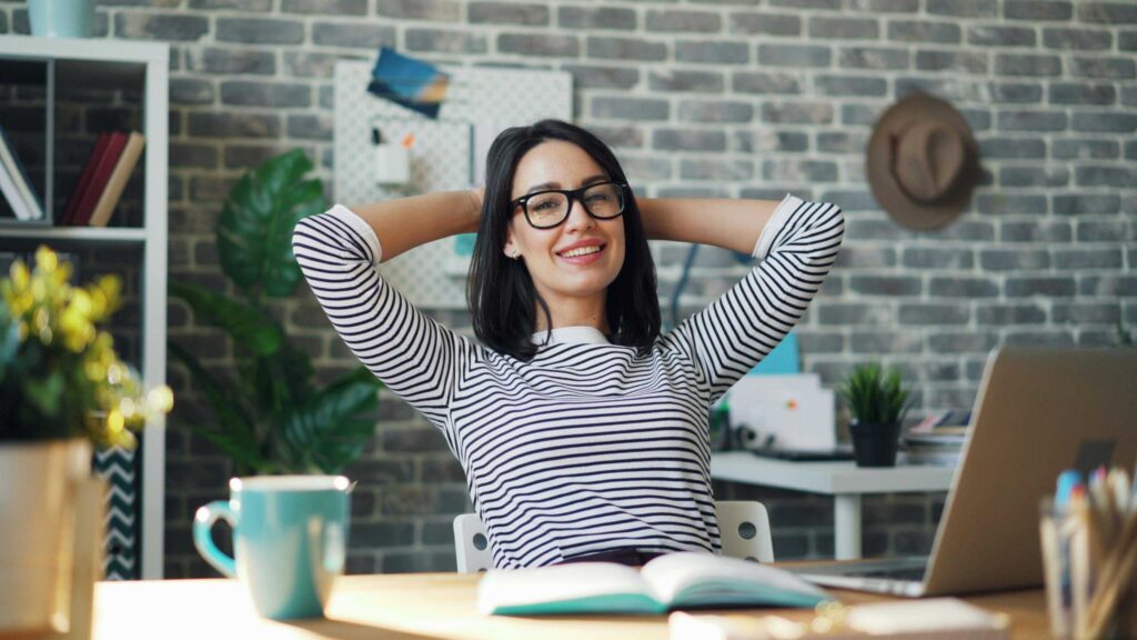 Smiling woman in a striped shirt and glasses sits at a desk with her hands behind her head in an office setting.