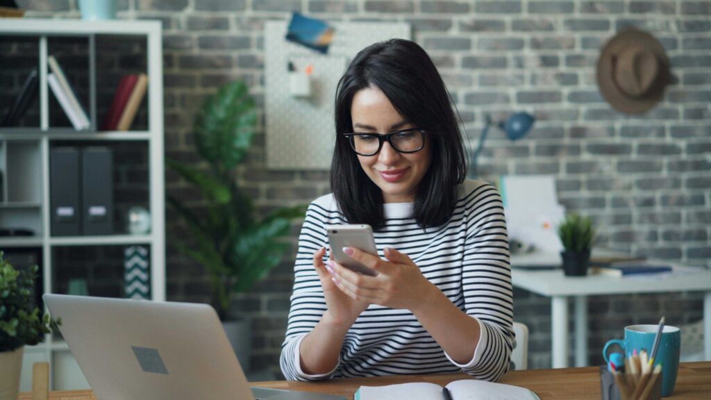 Woman with glasses sits at a desk in a striped shirt, looking at her smartphone with a laptop nearby in a modern office.