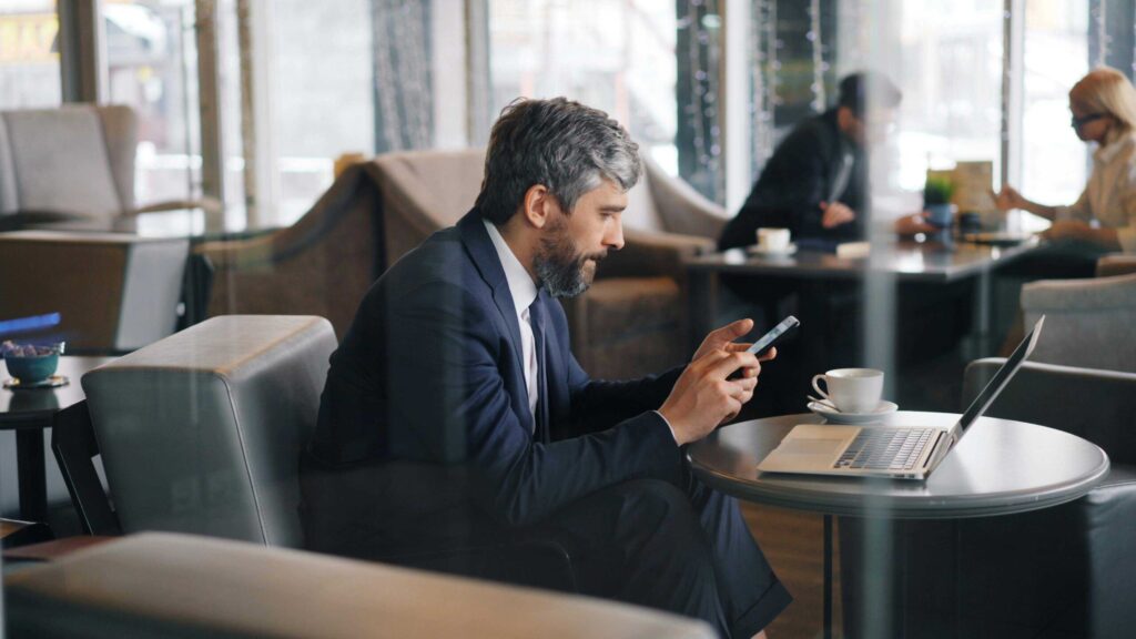 Man in a navy suit uses a smartphone at a cafe table with a laptop and coffee nearby