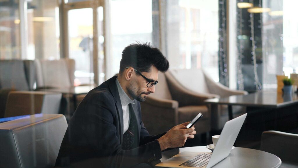 Professional man in a dark blazer using a smartphone while a laptop sits open on the table in a bright cafe lounge, coffee cup nearby