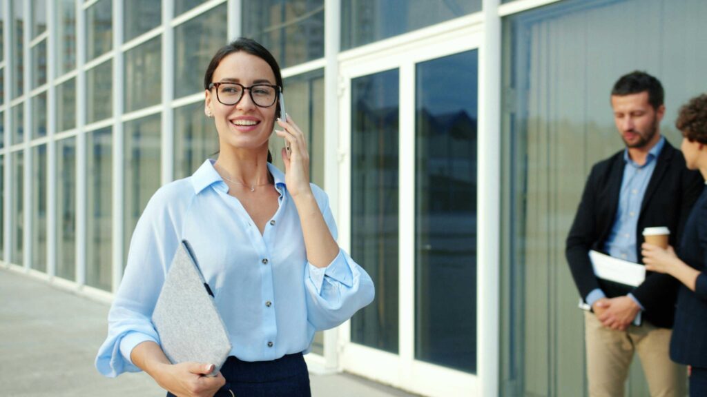 Smiling woman in a light blue blouse on a call outside a glass office building, holding a folder, with coworkers in the background.