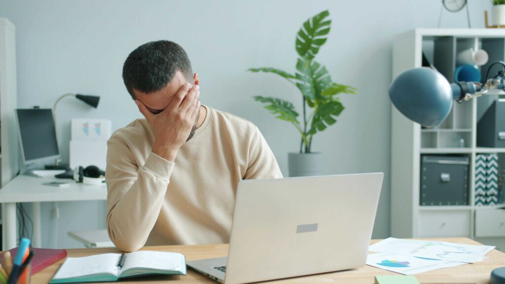 Man at a desk with laptop, head in his hand in a frustrated pose inside a bright office with a plant in the background.