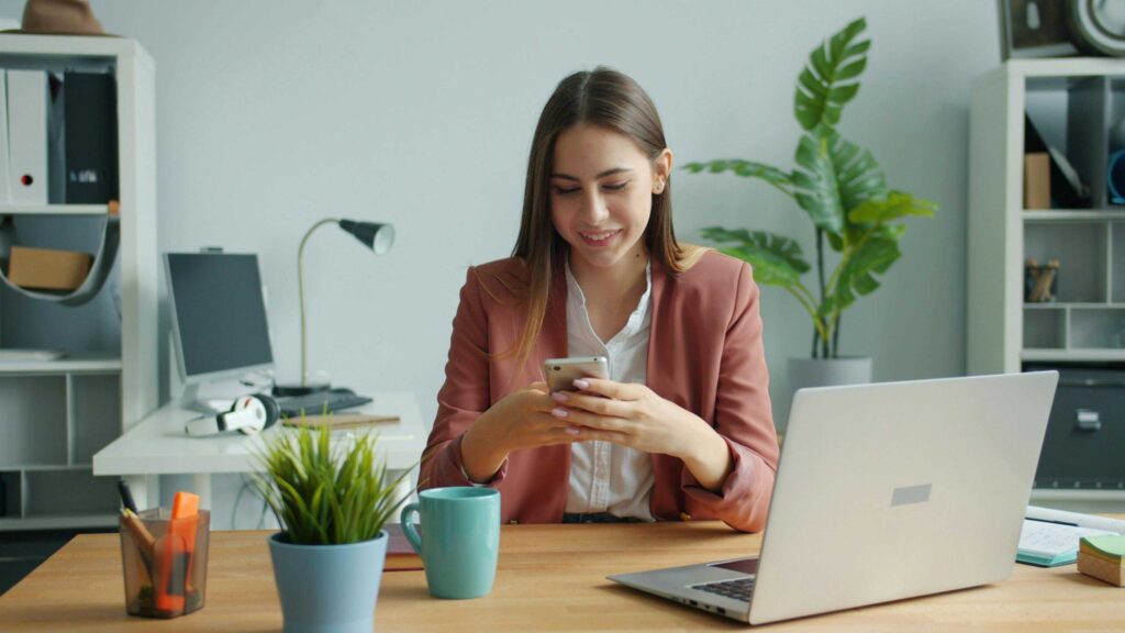Businesswoman in a pink blazer sits at a desk in a modern office, looking at her smartphone with a laptop nearby.