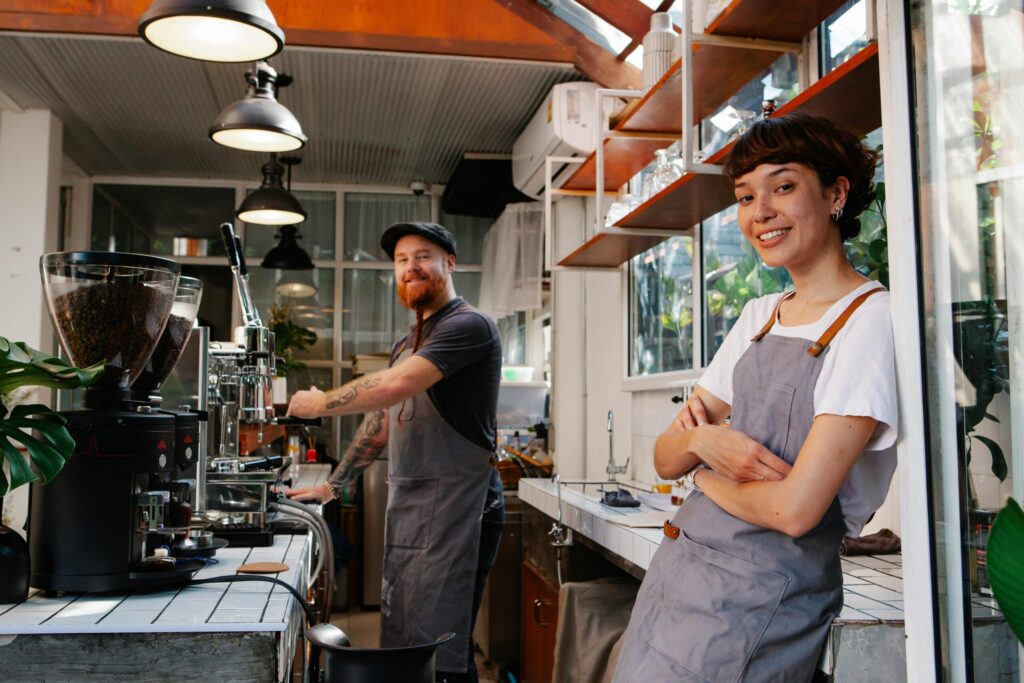 Smiling woman barista leaning at the cafe window in a gray apron, with a male barista and espresso machine in the background.