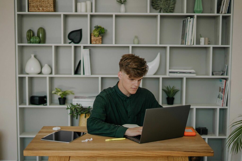 Man sits at a wooden desk typing on a laptop with white shelves and plants in the background