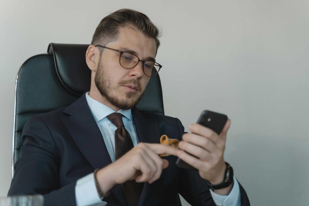 Man in a navy suit and brown tie, wearing glasses, sits in a black leather chair while looking at his smartphone with both hands visible to the camera.