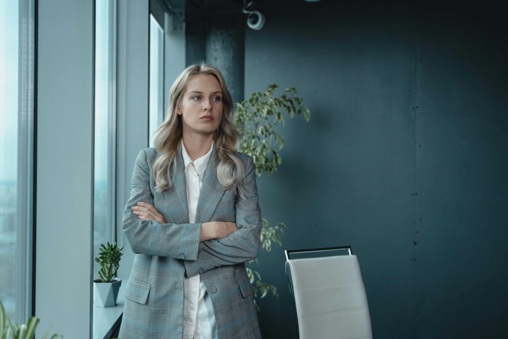 Businesswoman in a gray plaid blazer standing with arms crossed by a window in a modern office setting.