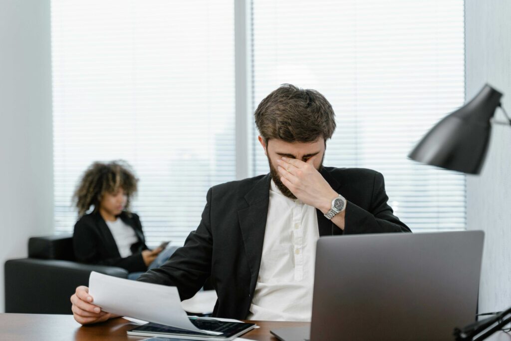 Bearded man in a suit at a desk, covering his face with his hand while holding papers, laptop nearby.