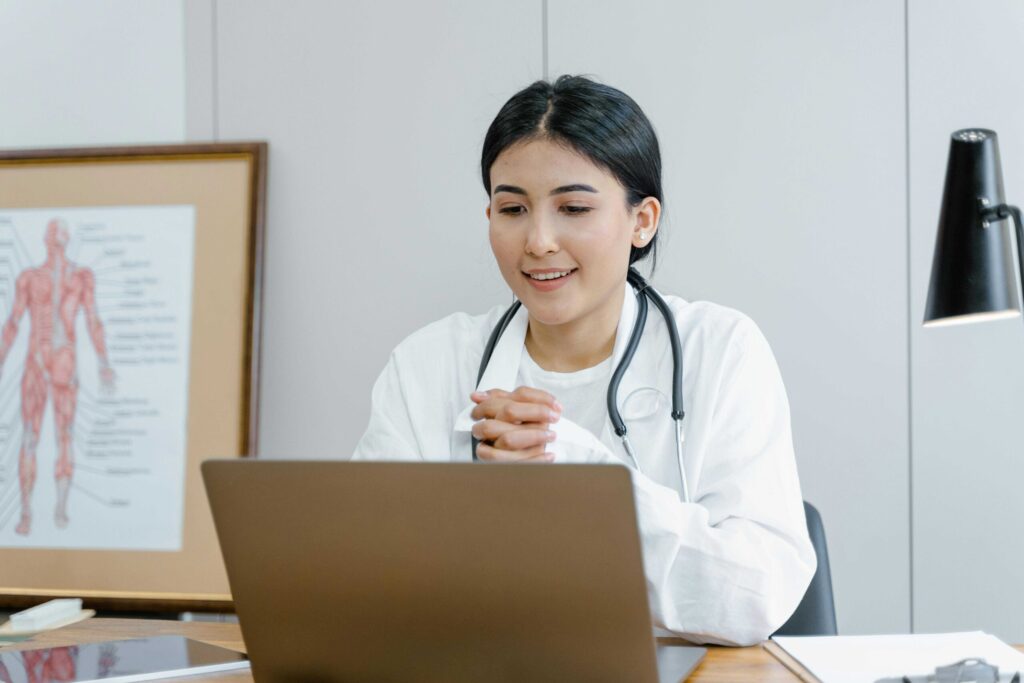 Female doctor in a white coat with a stethoscope sits at a desk, using a laptop in a clinic with a medical chart visible in the background.