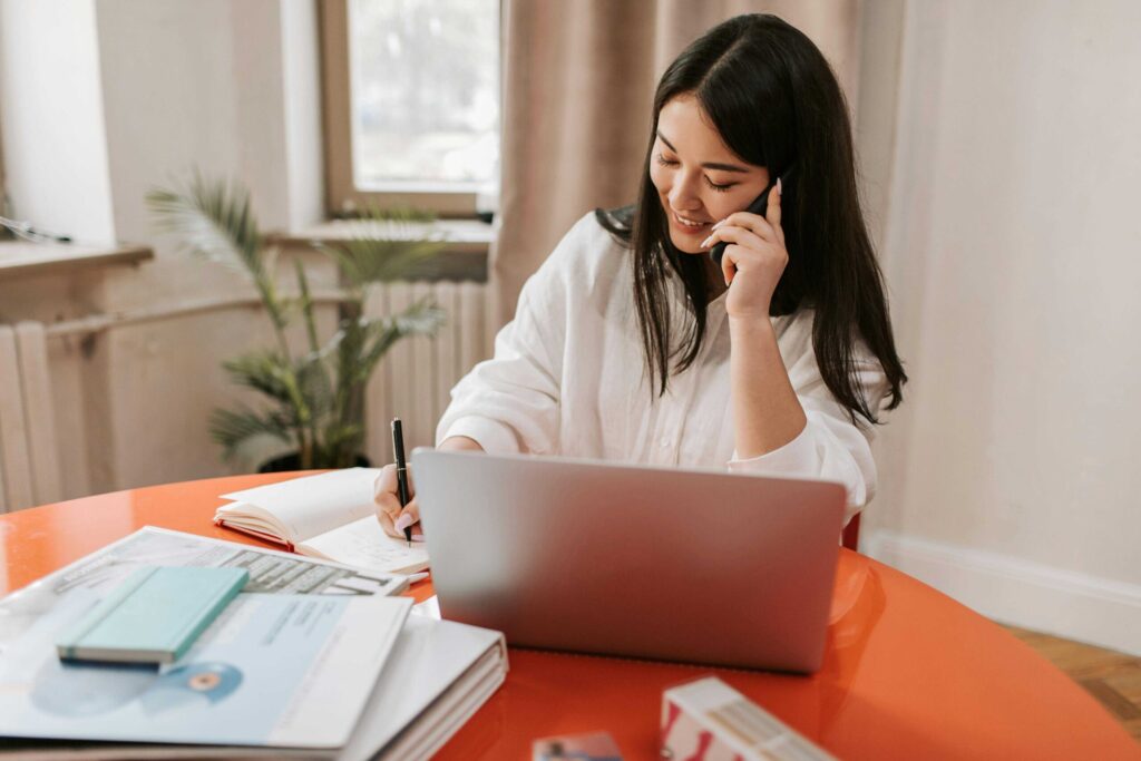 Woman smiling while talking on a phone, writing in a notebook at an orange table with a laptop in front of her in a bright home office.