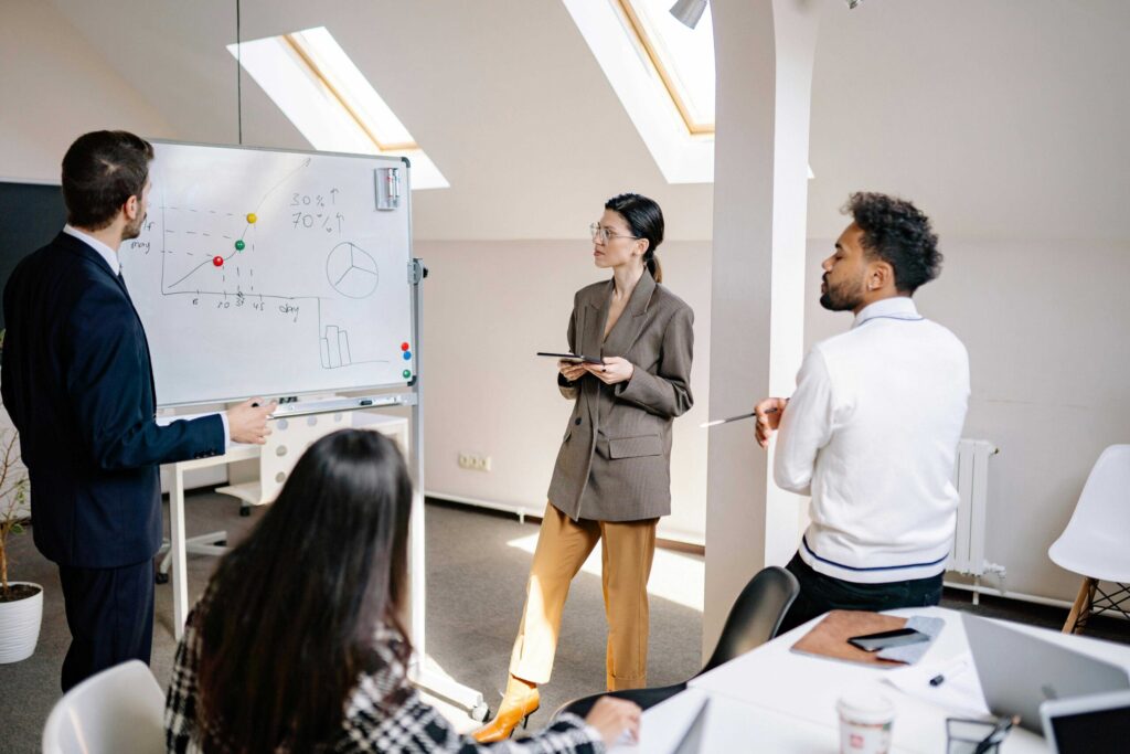 Group of professionals in a meeting room watching a presenter explain a graph on a whiteboard with colored markers.