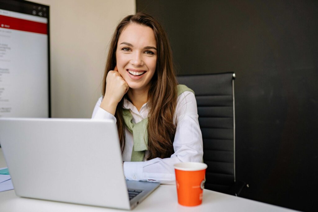 Smiling woman with long brown hair sits at a desk, resting her chin on her hand beside an open laptop.