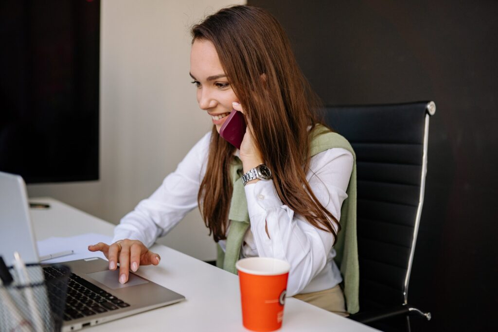 Woman seated at a desk, talking on a pink smartphone while typing on a laptop, red coffee cup in foreground.