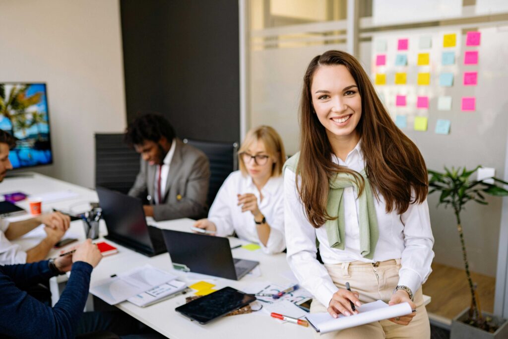 Smiling woman standing at a conference table while colleagues work on laptops in an office with sticky notes on a glass wall in the background.