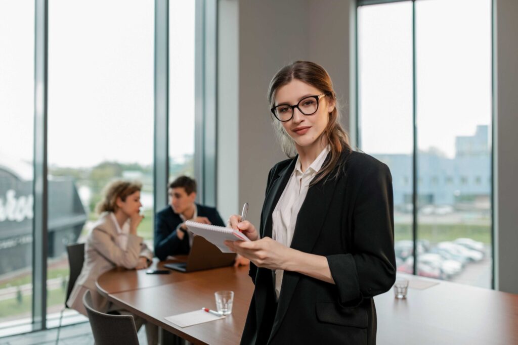 Businesswoman in a blazer stands at a conference table, holding a notebook and pen while colleagues discuss in the background.