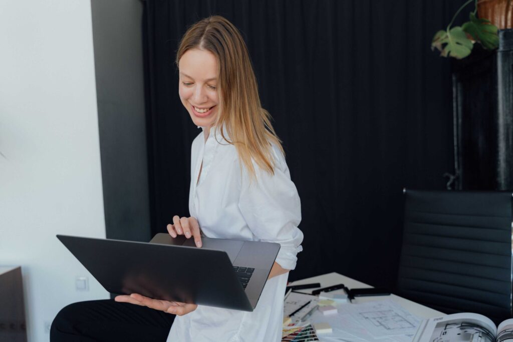 Woman in a white shirt standing and smiling while using a laptop in an office setting.