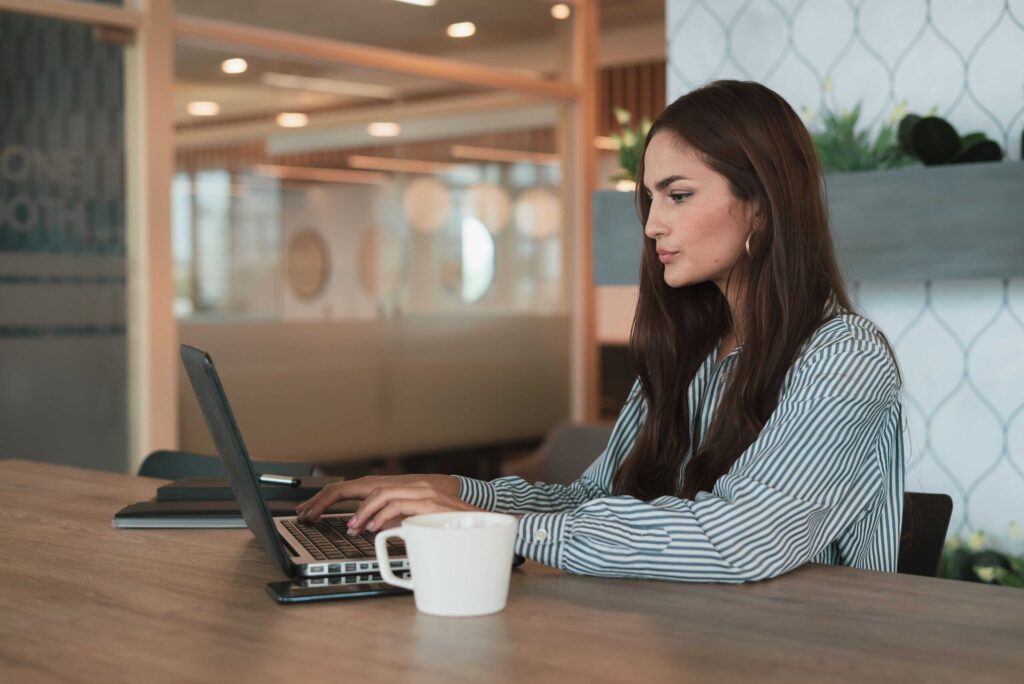Woman in a striped shirt typing on a laptop at a wooden table in a modern office, with a white coffee mug nearby.