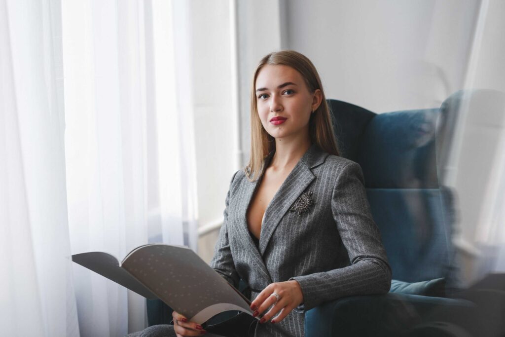 Professional woman in a gray pinstripe blazer seated by a window, reading a magazine.