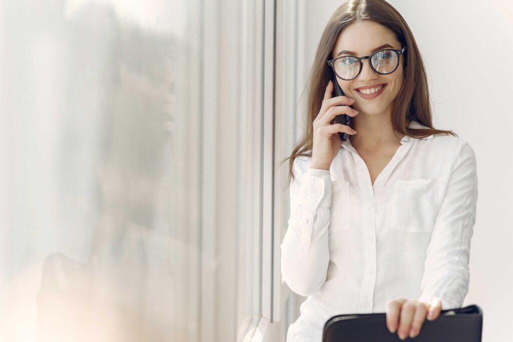 Smiling woman with glasses in a white blouse talks on a mobile phone, holding a black briefcase in a bright office setting.