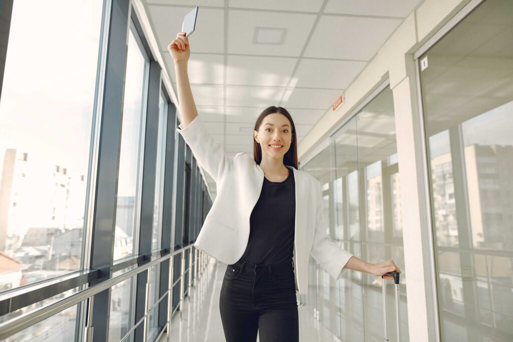 Smiling woman in a white blazer raises a passport in the air while walking with a rolling suitcase in a glass-walled hallway.