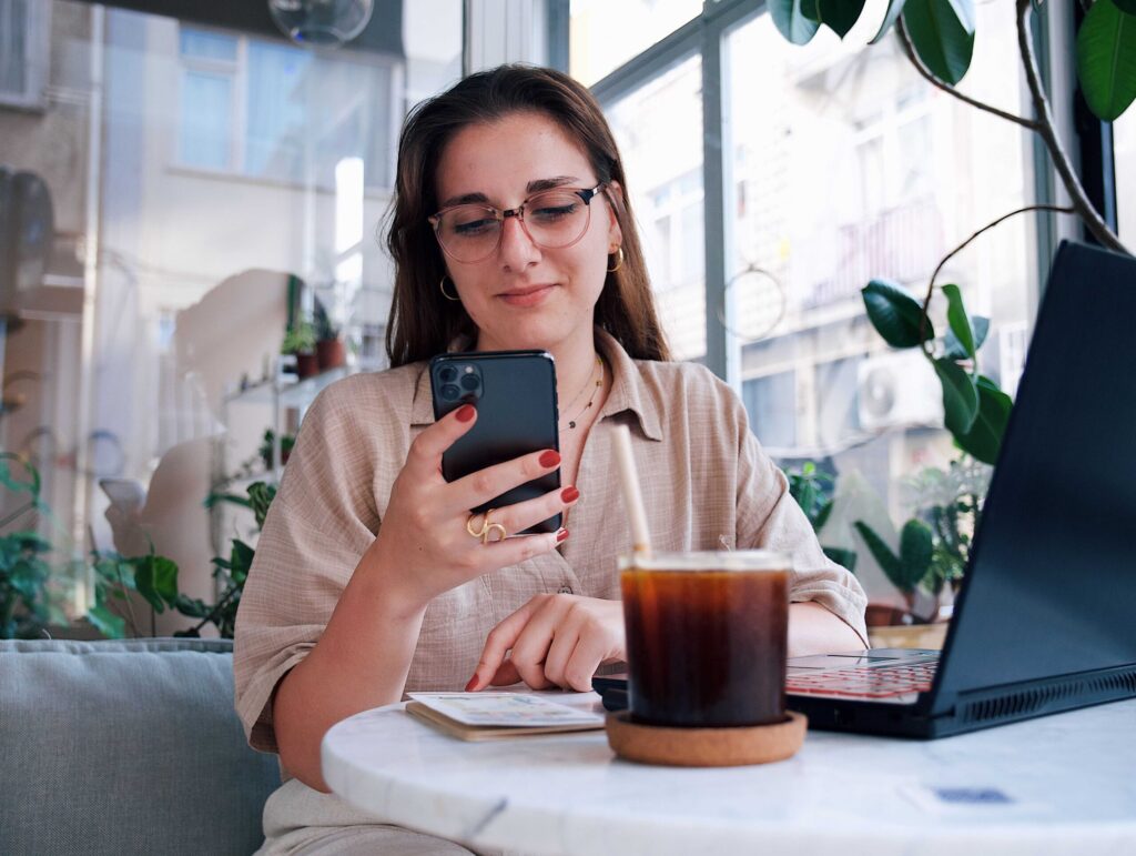 Woman wearing glasses at a cafe table, looking at her smartphone with a laptop and iced coffee nearby.